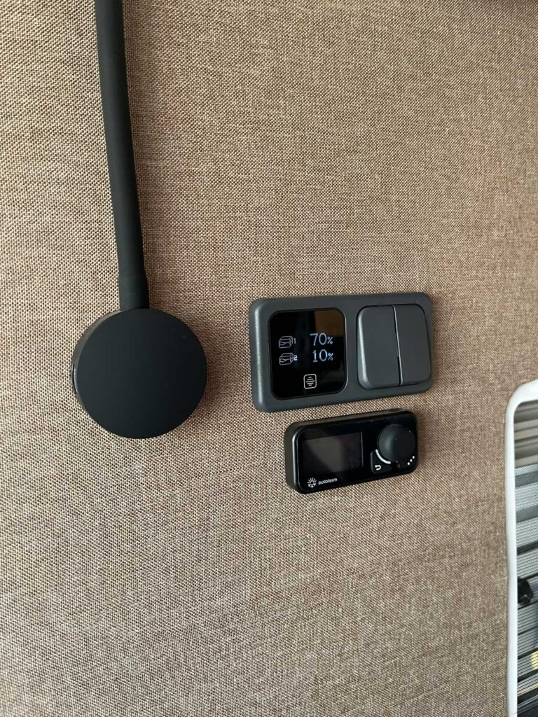 Un primer plano de una pared beige con un dispositivo electrónico, dos interruptores grises que muestran 70% y 10°C, y un dispositivo circular negro montado sobre un pequeño panel de control digital-ideal para la camperización de furgonetas en Castellón.