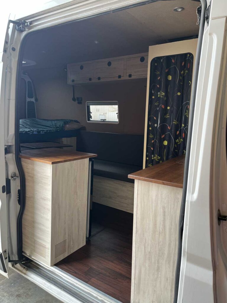 Vista del interior de una furgoneta camper con armarios de madera, un banco acolchado y espacio de almacenamiento en la parte superior: un ejemplo de camperización de furgonetas en Castellón que crea un espacio de vida acogedor y organizado. La puerta de la furgoneta está abierta, dejando entrar la luz natural.
