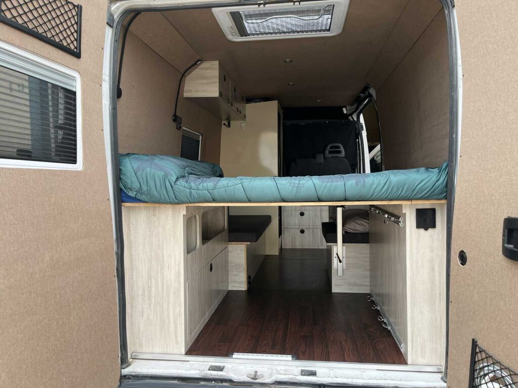 La foto muestra el interior de una furgoneta camper, expertamente transformada mediante camperización de furgonetas en Castellón, con una cama elevada con almacenaje debajo, suelo de madera, armarios y estanterías a lo largo de las paredes.