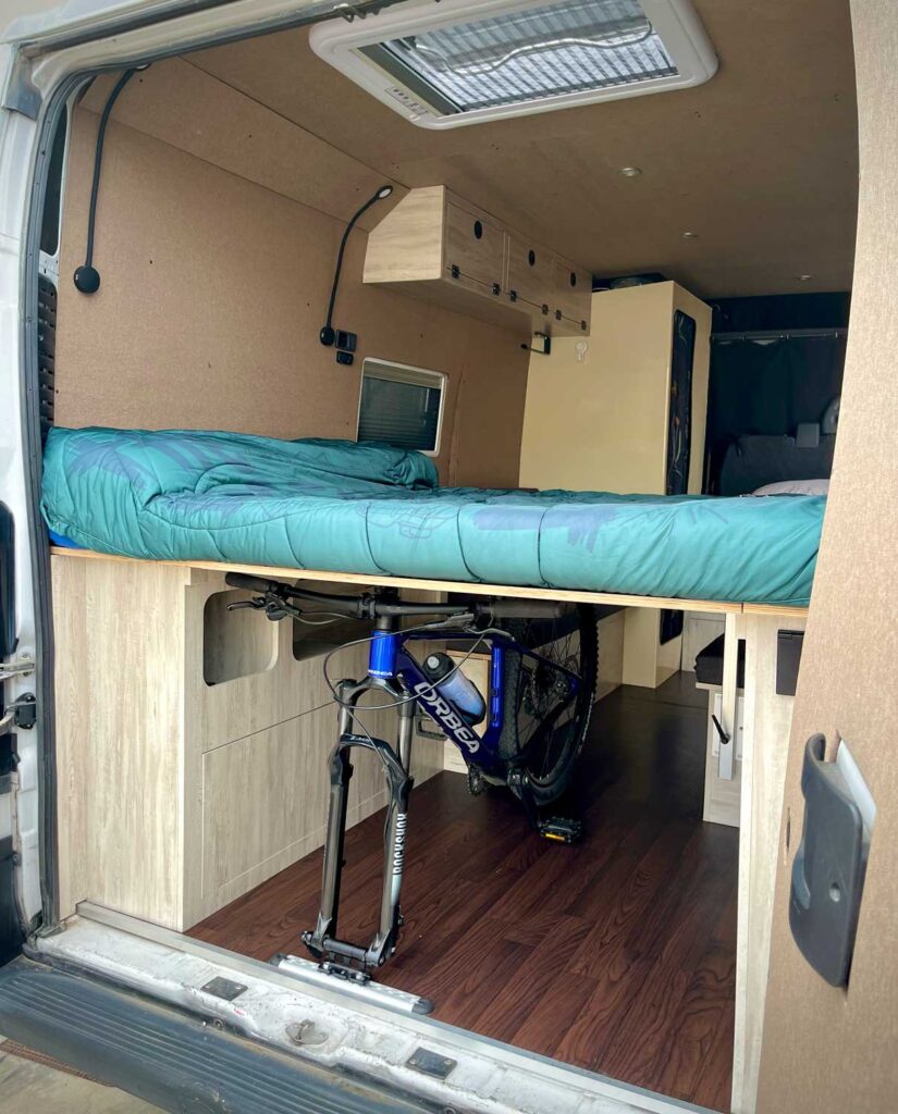 Un interior de furgoneta camper diseñado para la camperización de furgonetas en Castellón, con una cama elevada con cajones de almacenamiento, una bicicleta de montaña montada debajo, paneles de madera, armarios y una rejilla de ventilación en el techo para mayor comodidad y conveniencia.