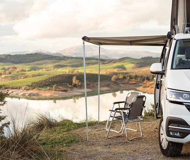 Una furgoneta blanca expertamente adaptada con camperización de furgonetas descansa junto a un lago. Dos sillas vacías bajo su toldo extendido dominan las verdes colinas onduladas y el agua resplandeciente bajo un cielo nublado.