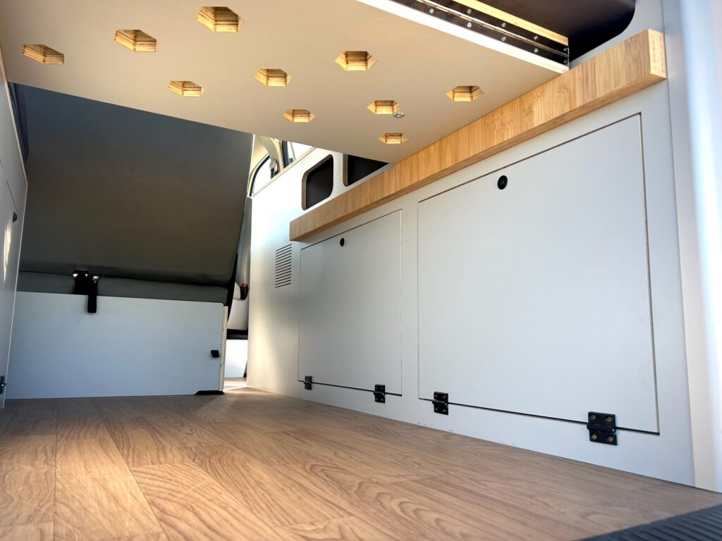 Vista del interior de una moderna furgoneta camper tras una elegante camperización de furgonetas, con suelo de madera, armarios blancos con bisagras negras, encimera de madera y lámparas hexagonales en el techo. El espacio es limpio y minimalista.