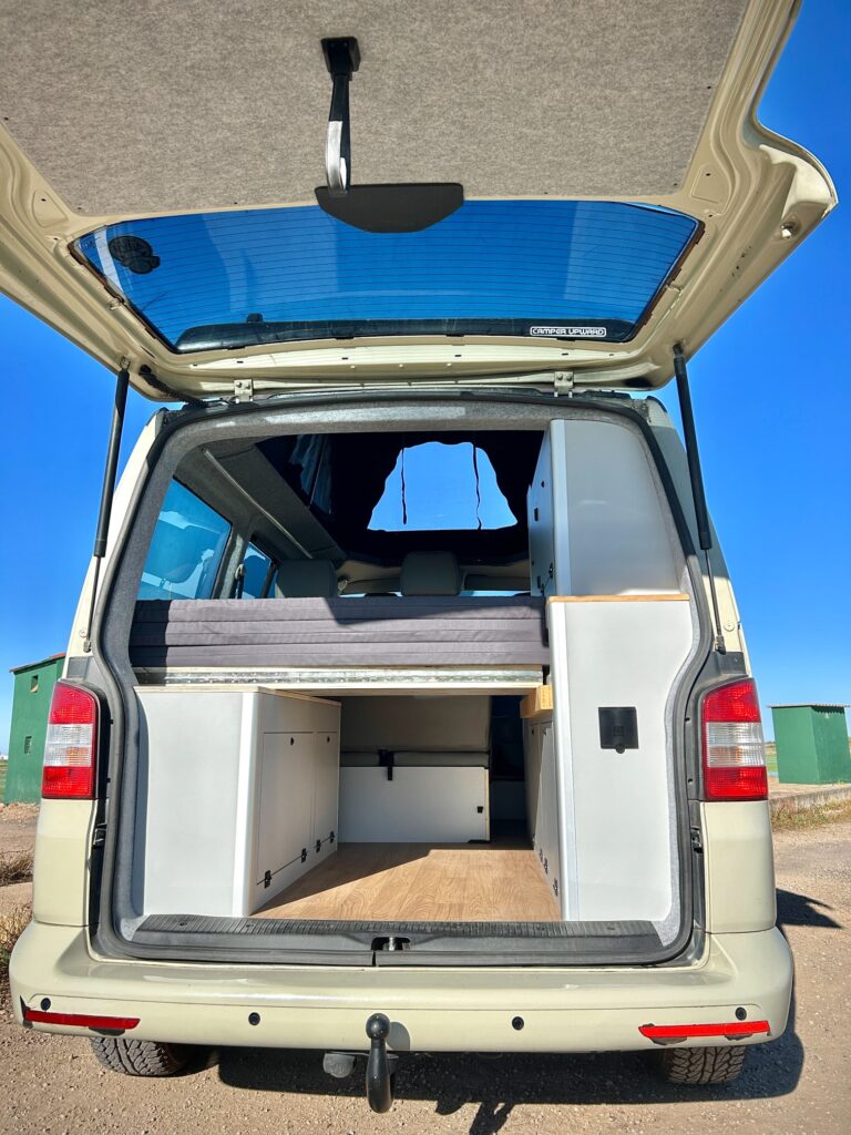 La vista trasera de una furgoneta camper con la puerta trasera abierta revela una elegante camperización de furgonetas, mostrando un interior compacto y moderno con almacenamiento integrado, armarios, una plataforma de cama y suelos de madera bajo un cielo azul despejado.