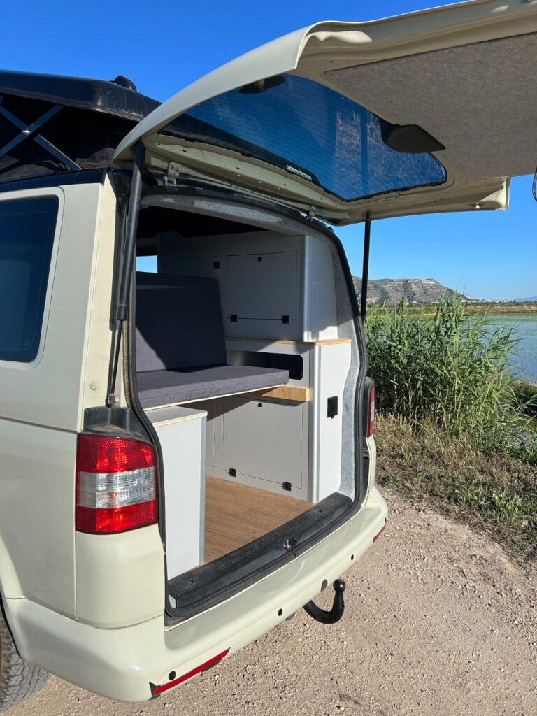 Una furgoneta camper con la puerta trasera abierta, revelando un interior compacto de camperización de furgonetas con asientos, armarios y suelo de madera, aparcada cerca de un río con juncos y colinas al fondo bajo un cielo azul despejado.