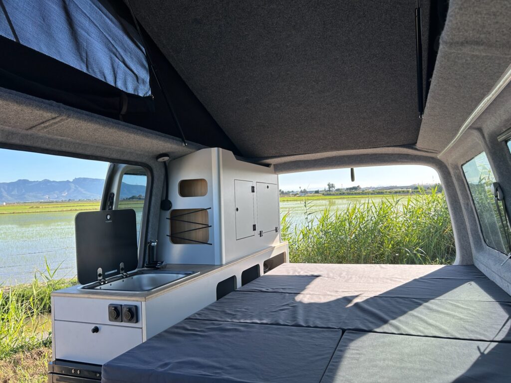 Interior de una furgoneta camperizada con techo elevado, mostrando una compacta cocina con fregadero y fogones, armarios y cama plegada. Grandes ventanales permiten disfrutar del paisaje verde y de un campo exterior. Ideal para la camperización de furgonetas.
