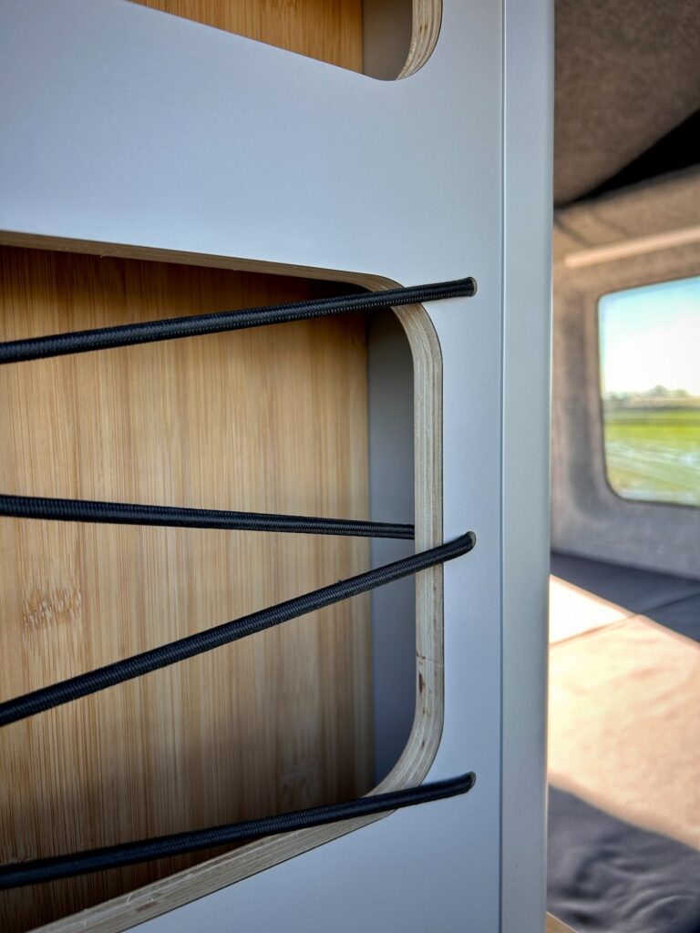 Primer plano de una estantería empotrada en la furgoneta con cordones elásticos negros y un panel de madera, la luz del sol entrando por una ventana y el verde paisaje exterior: un detalle perfecto en la camperización de furgonetas para viajar de forma organizada y con estilo.