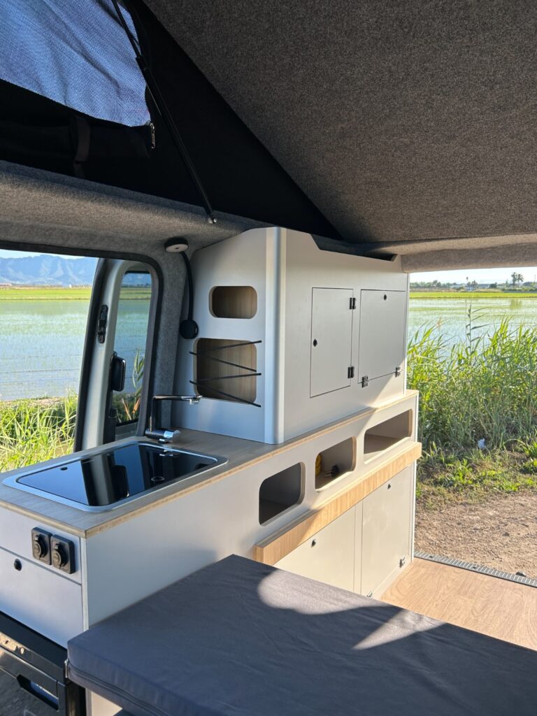 El interior de una moderna furgoneta camper muestra una experta camperización de furgonetas, con una pequeña cocina, fregadero, fogones, armarios y una cama. La puerta abierta de la furgoneta enmarca vistas de hierba verde, agua y montañas lejanas.