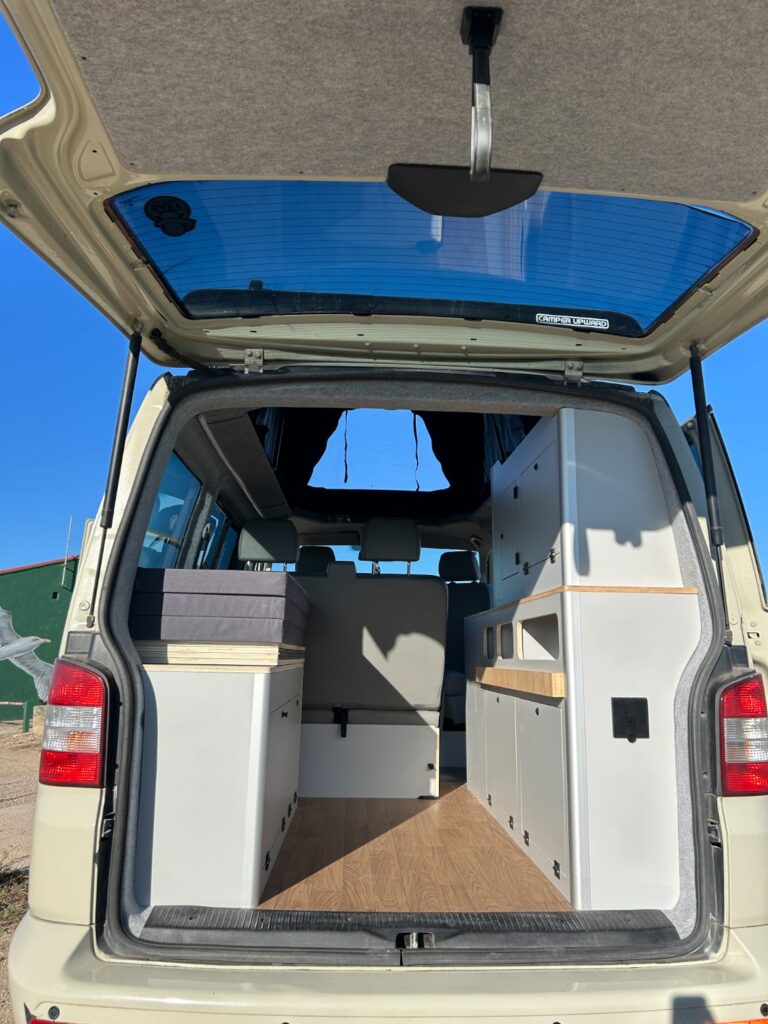 La foto muestra la parte trasera abierta de una furgoneta camper beige, revelando un interior pulcro y moderno con armarios blancos y superficies de madera -un inspirador ejemplo de camperización de furgonetas- bajo un cielo azul despejado.