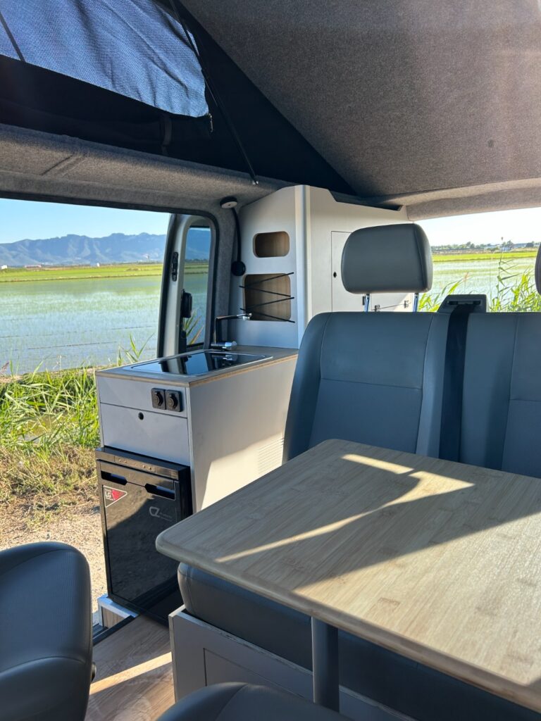 Interior de una camperización de furgonetas: pequeña cocina, nevera y mesa plegable. Asientos grises frente a la mesa. Grandes ventanales muestran un paisaje con agua, césped verde y montañas a lo lejos.