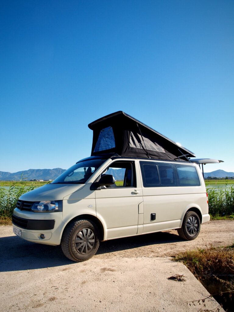Una furgoneta camper blanca con techo desplegable, resultado de experta camperización de furgonetas, está aparcada en un camino de tierra rodeado de hierba alta, con montañas y un cielo azul despejado al fondo.