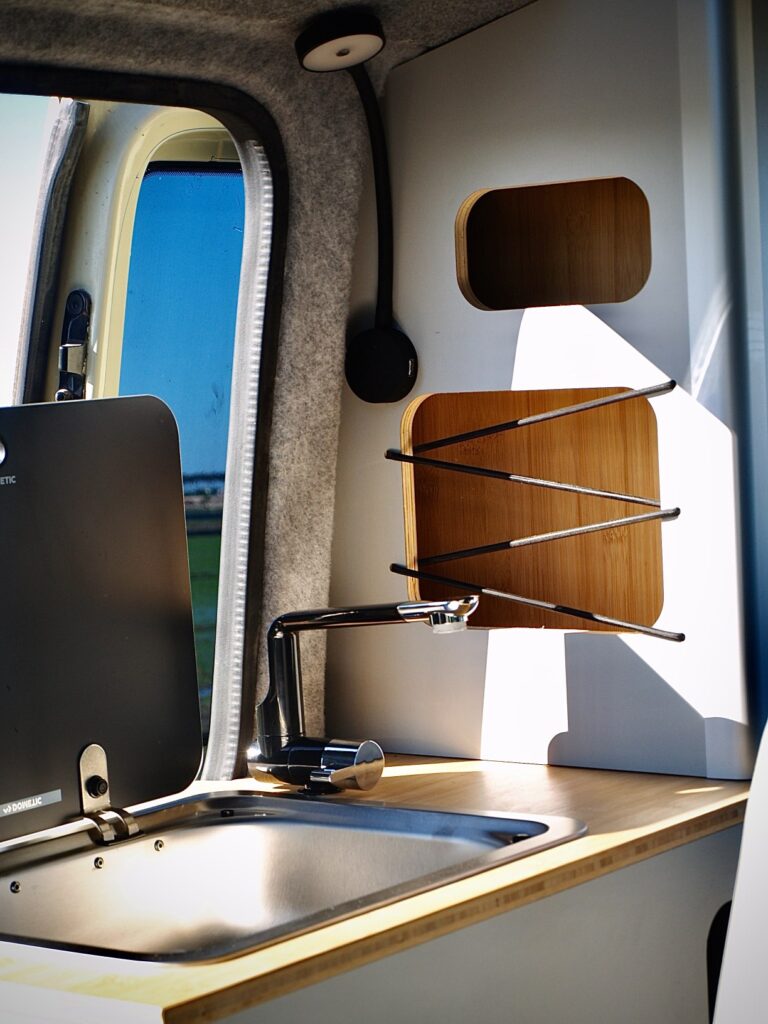 Una moderna cocina de furgoneta camper exhibe una experta camperización de furgonetas, con un fregadero de acero inoxidable, un grifo elegante, una tapa de cocina negra cerrada y estantes de madera con barras metálicas junto a una ventana iluminada por el sol.