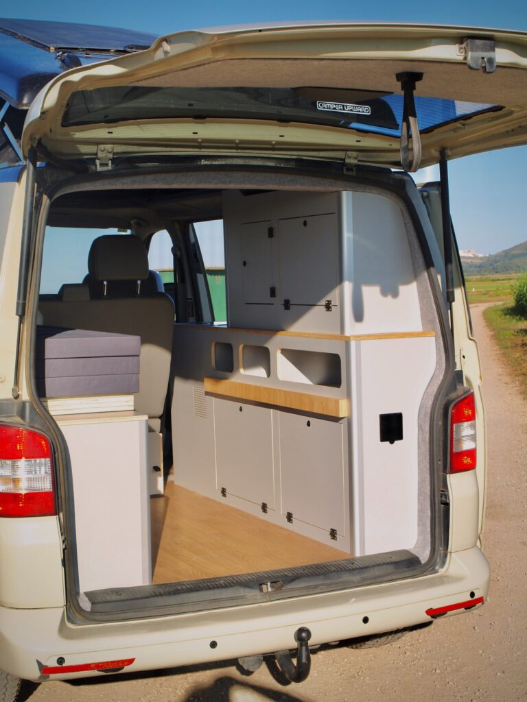 Una vista de la parte trasera de una furgoneta camper con la puerta trasera abierta revela una elegante camperización de furgonetas, con armarios blancos empotrados, estanterías y una zona de asientos acolchados sobre un suelo de madera, aparcada en un camino de tierra junto a zonas verdes.