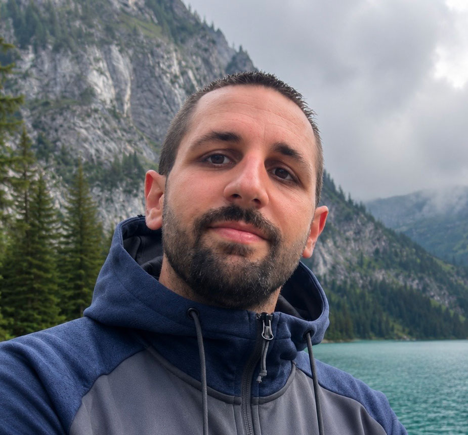 Un hombre de pelo corto y oscuro y barba, con una chaqueta azul con capucha, está de pie frente a un lago con pinos y montañas rocosas -un lugar perfecto para quienes disfrutan de la camperización de furgonetas- en un día nublado.